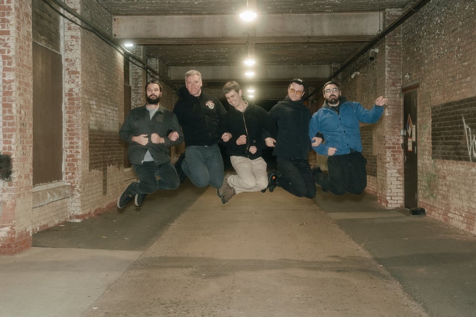 Five men in a line are photographed in mid-jump in a brick-lined alleyway.