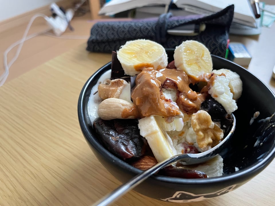 An indulgent bowl of ice cream that is so covered in toppings you can barely see the ice cream below it. Visible from the picture are slices of banana, a peanut butter drizzle, cashews, walnuts, and dates.