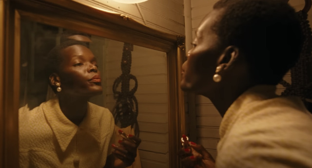 [A woman in a cream blouse and short cropped hair holds a lipstick and purses her lips in the mirror, inspecting its application.]