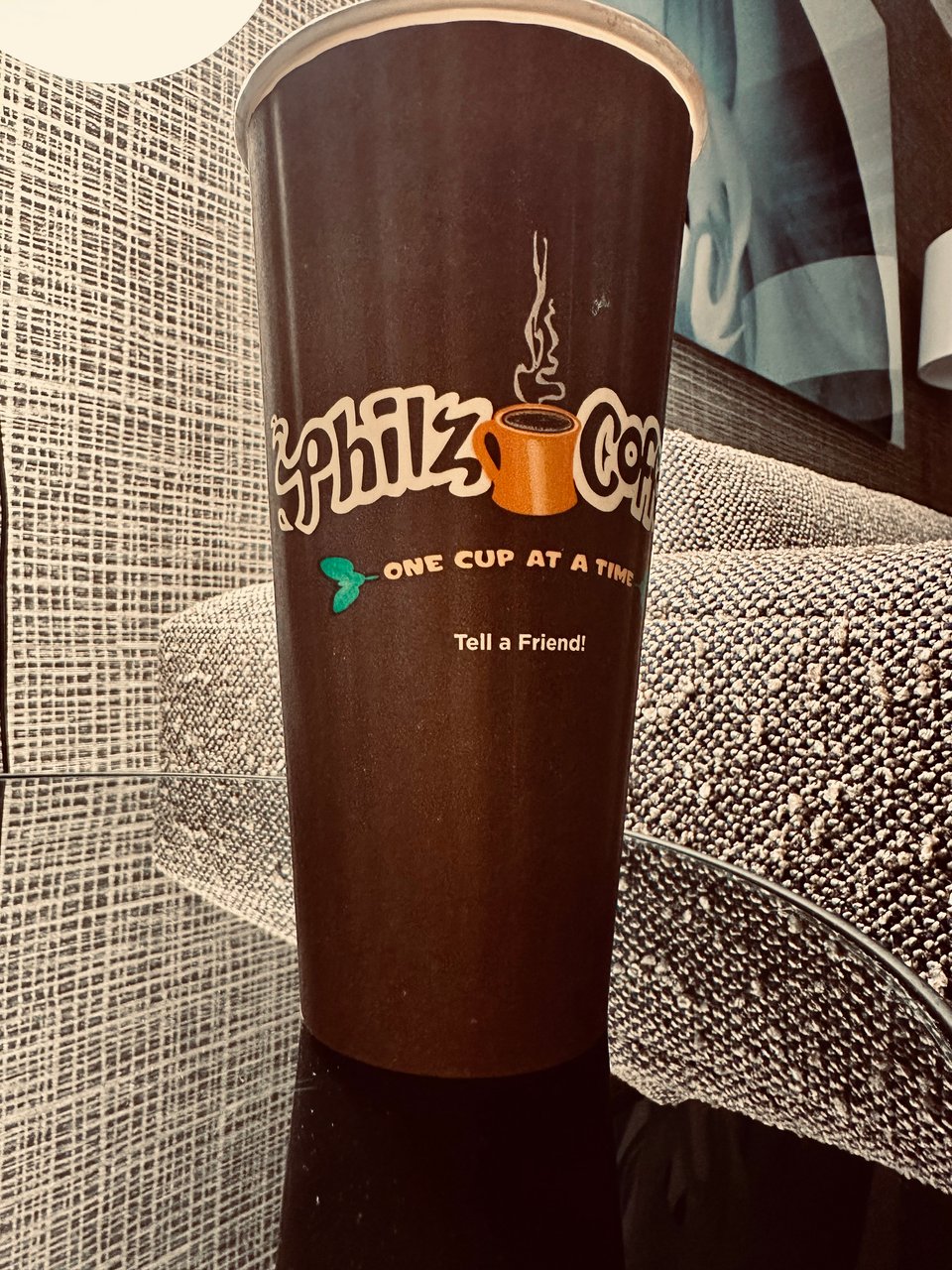 photo of a large cardboard cup of coffee on a black reflective hotel nightstand, beside a sofa with knit upholstery