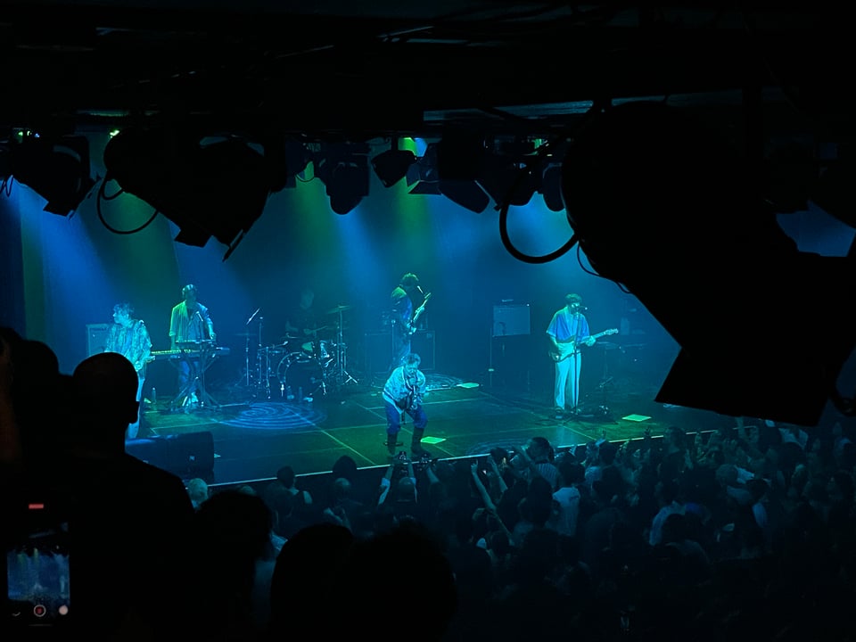 Concert hall with the band on the stage and everyone bathed in green and blue light.