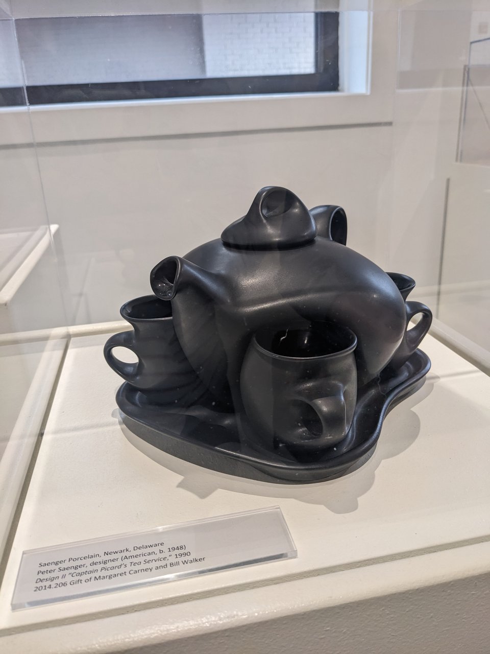A black tea set in a glass vitrine. The mugs are molded to fit around the kettle.