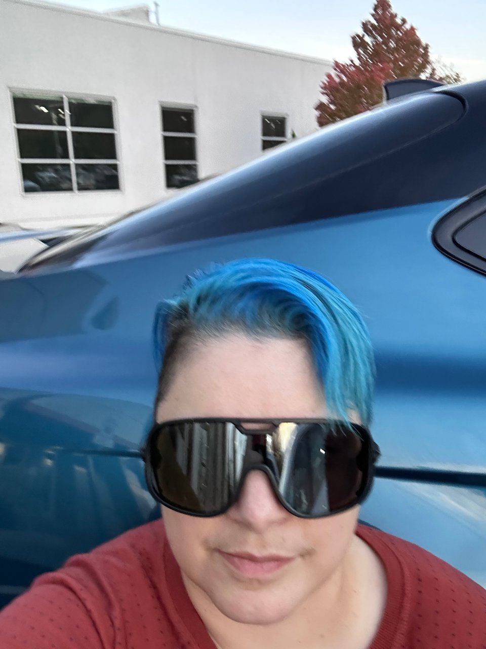 A white person with dark wraparound sunglasses and messy short teal hair crouches in front of shiny new teal car.