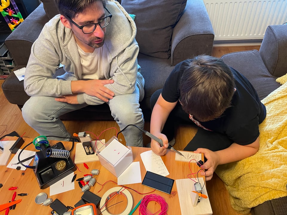 A man sits next to a boy in the living room explaining as the boy used a soldering iron to burn his name into a piece of balsa wood.
