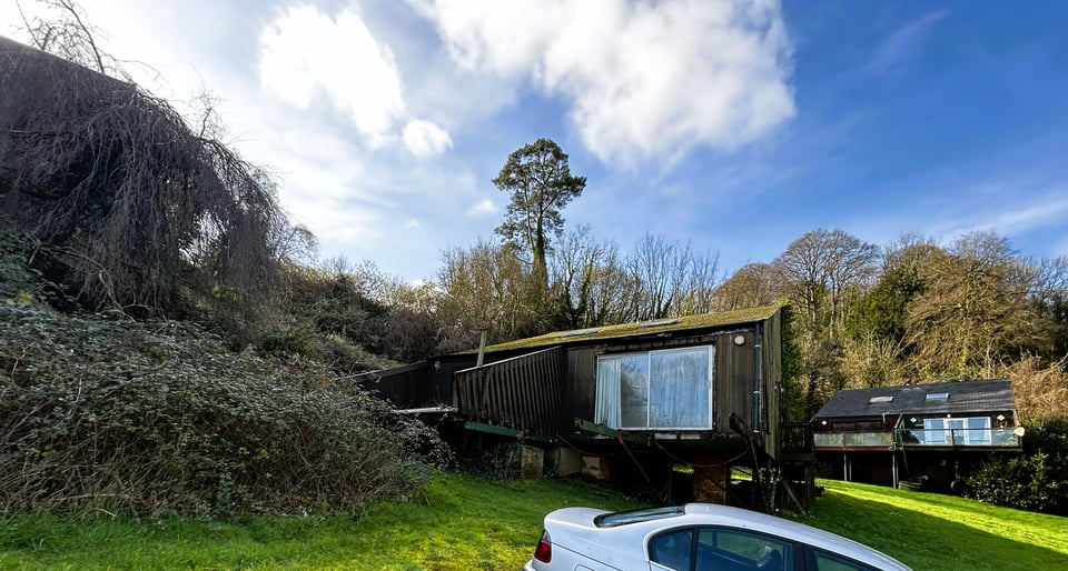 The second three pairs of lodges from 1979. In the background is a complete lodge. It's balconies are supported by props but they are otherwise OK. The middle pair are empty, the one on the left is rotting and the one on the right is missing its balcony and has steel beams sticking out instead. In the foreground, brambles and climbers have almost entirely obscured the last of the three. The woods beyond are sunlit gold against a blue sky.