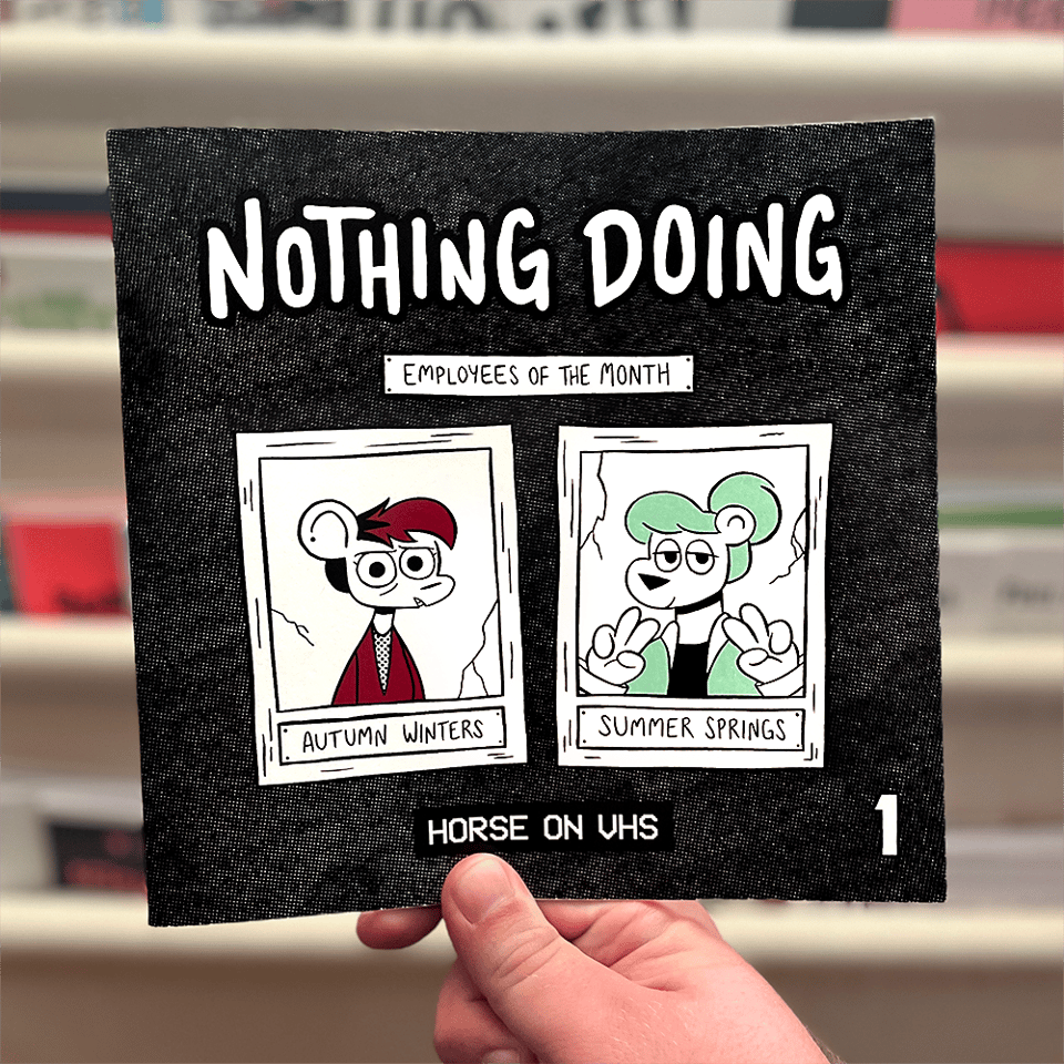 a photo of a hand holding up Nothing Doing #1. the cover features Autumn Winters and Summer Springs in cracked and crooked "employee of the month" portraits with their names under them, the former looking concerned and the latter throwing dual peace signs.