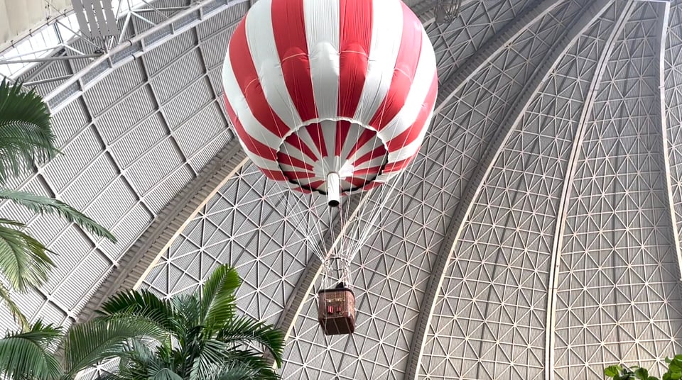 A view from below of a red and white striped balloon with a whicker basket hanging from it. Above it is the dome, below are the fringes of palm trees.