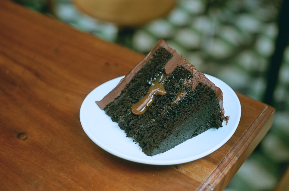 A slice of dark chocolate cake with light chocolate frosting and a drip of caramel sits on a white plate on the corner of a brown wooden table, above a blurred-out tile floor