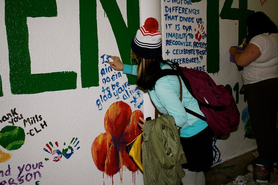 Jen is painting with a finger on a wall that also has a lot of hand painted quotes on it. In blue paint, Jen has written, "It ain't no sin to be glad your'e alive."
