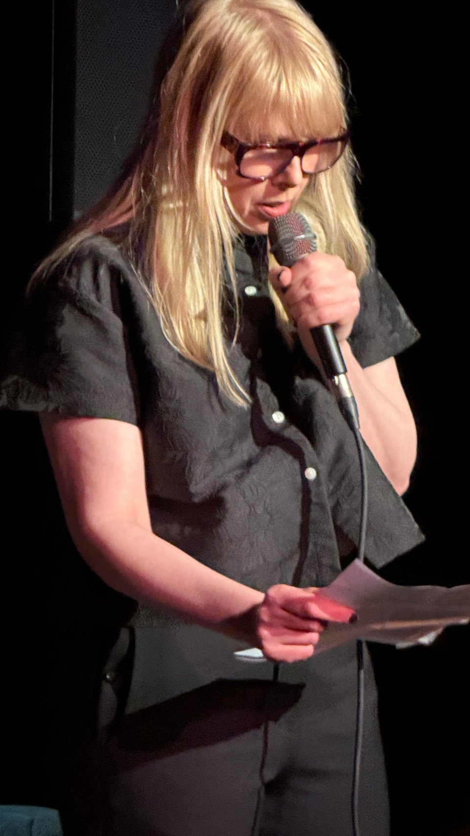 Johanna Fateman, three-quarter view, wearing black and heavy reading glasses spotlit in a dark room , clutching a microphone.