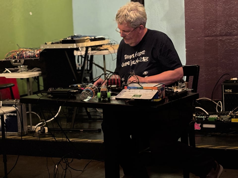 S Glass performing live sitting a table covered in electronic instruments