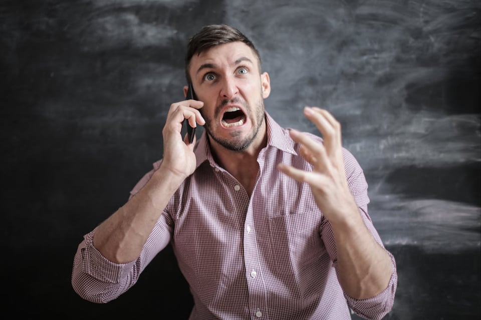 A picture of a man with short black straight hair and a red checked shirt; he is yelling angrily into a phone held to his ear, and gesticulating with his other hand