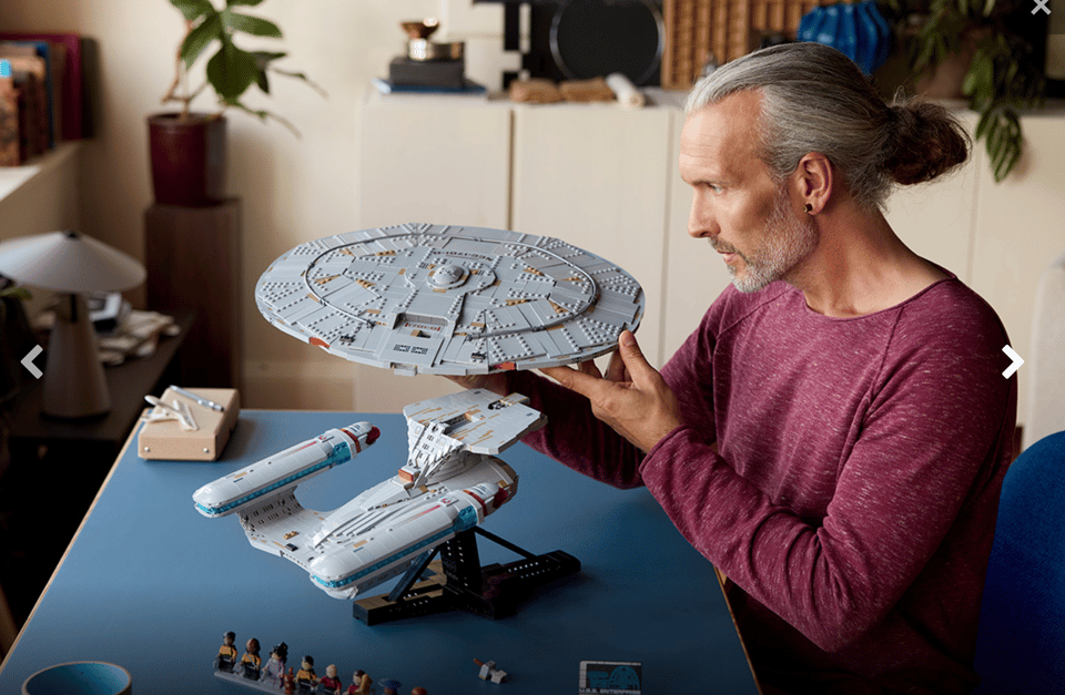 Photo of white man with long hair tied back and a well-trimmed beard, assembling & contemplating a Lego version of the starship Enterprise