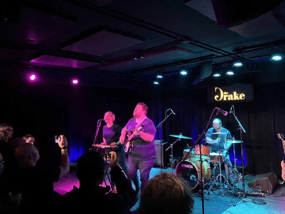 A woman on keyboards sings into a microphone next to a man playing guitar. Behind them is a man on drums.