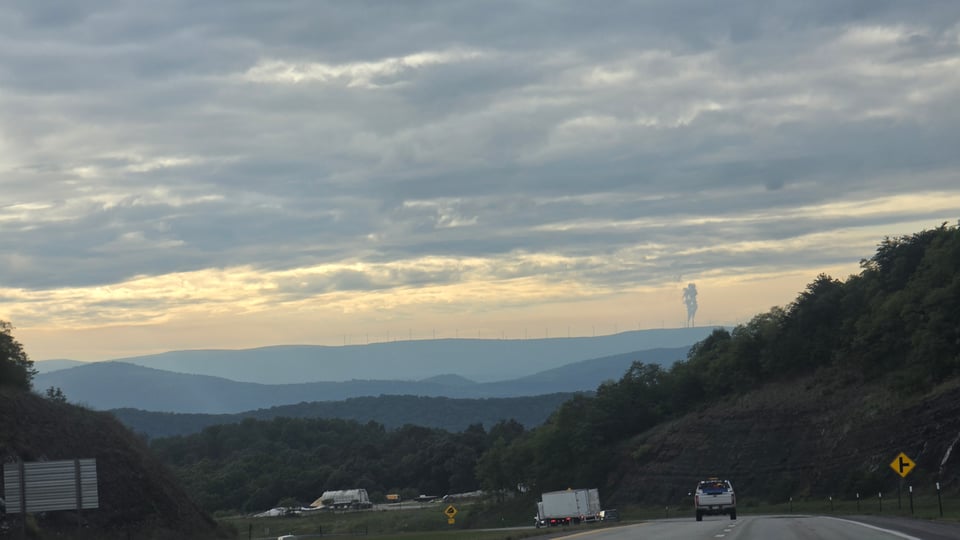 A highway winds through forested hills at dusk, with a white box truck and a pickup truck traveling ahead, distant wind turbines lining a far ridge beneath a broad, cloud-streaked sky tinted gold near the horizon.