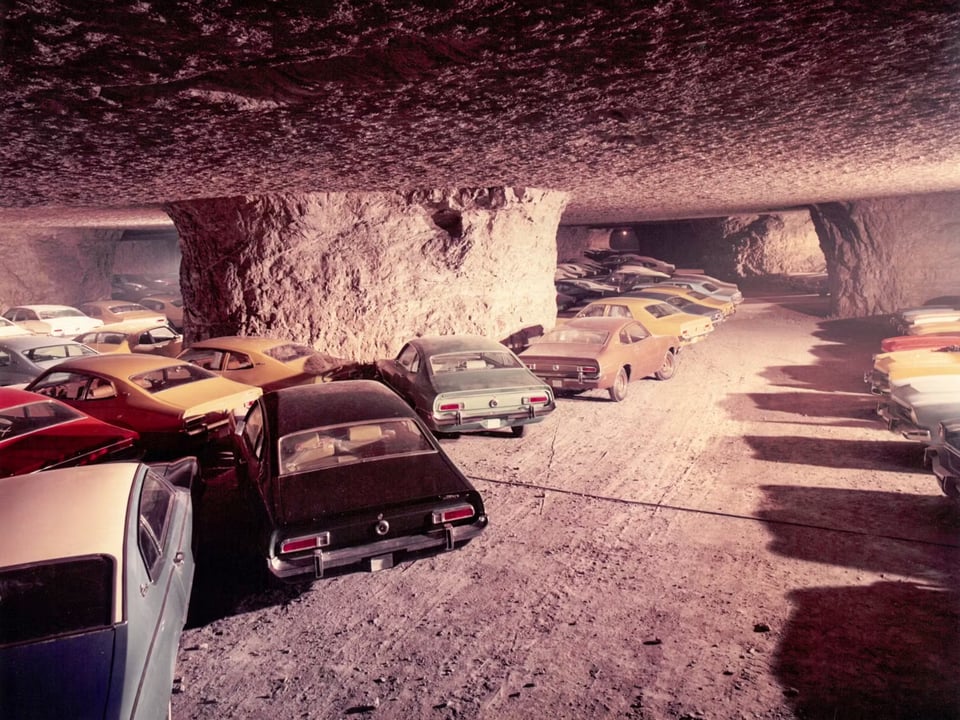 A picture of several 1960s and 1970s-era cars parked in a cave. The ceiling is low, the floor and walls and ceiling are all the same jagged stone material. The photo is slightly sepia-toned and it is clear it's from the 70s.