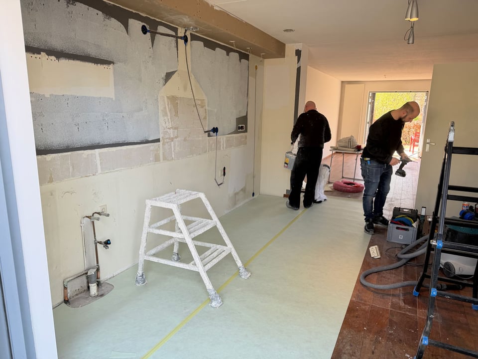 Two men work on a kitchen renovation which has been stripped to its bare bones, one an electrician and the other the plasterer.