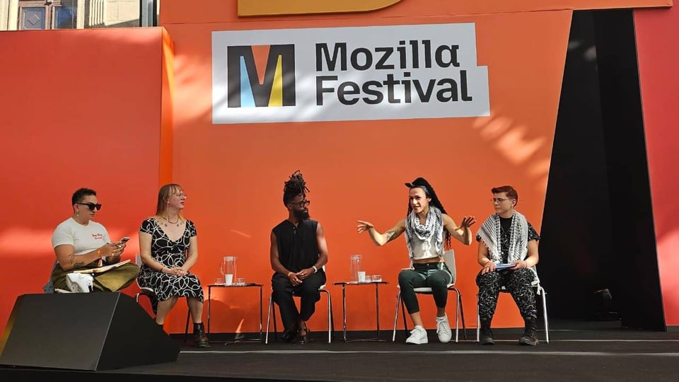 Unthinking Cisnormativity at Mozfest. From left to right: Alex Hanna (brown trans woman wearing sunglasses with a white t-shirt, short cropped black hair), Sasha Costanza-Chock (white trans woman wearing glasses and a black and white patterned dress, shoulder length blonde hair), Elijah McKinnon (Black non-binary person with locks atop their head, wearing a flowy black outfit), Veronyka Gimenes (Brazilian travesti non-binary person, shoulder length hair, wearing jeans, a crop top, and a Palestinian kuffiyeh), and Dia Kayyali (white-presenting non-binary person with short red hair, wearing Palestinian kuffiyeh and dark pants). Speakers are all fabulous trans people sitting on a stage with an orange background. Above it, it reads Mozilla Festival.