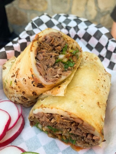 A burrito filled with birria and onions sits propped up in a basket with a black and white checked liner