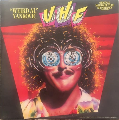 A picture of Weird Al from the shoulders up. He has a long narrow head, a brown mustache, and a big head of curly brown hair. He's wearing a Hawaiian shirt. His eyes are replaced with blue grey TV test patterns. He has a slight grin on his face. The sky behind him is red.
