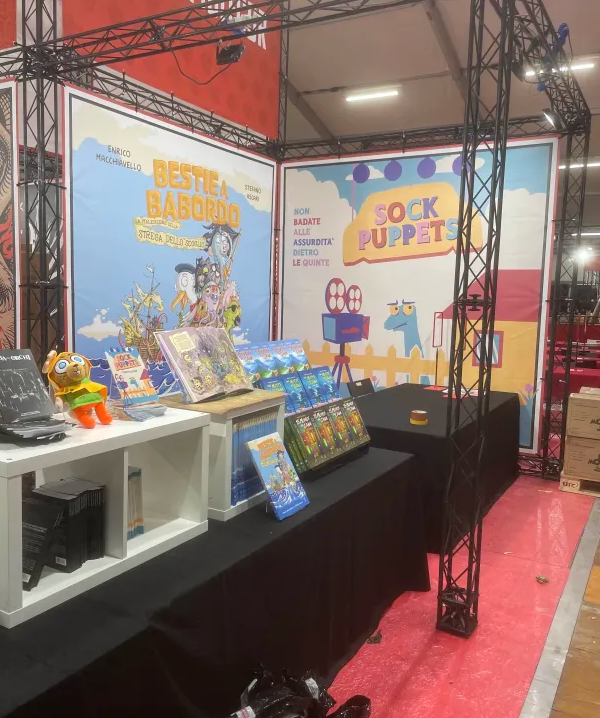 A convention booth. The back wall of the booth is a banner image promoting Sock Puppets, and it's enormous: perhaps 7 by 7 feet. In the foreground, the book can be seen on a stand, with copies for sale underneath.