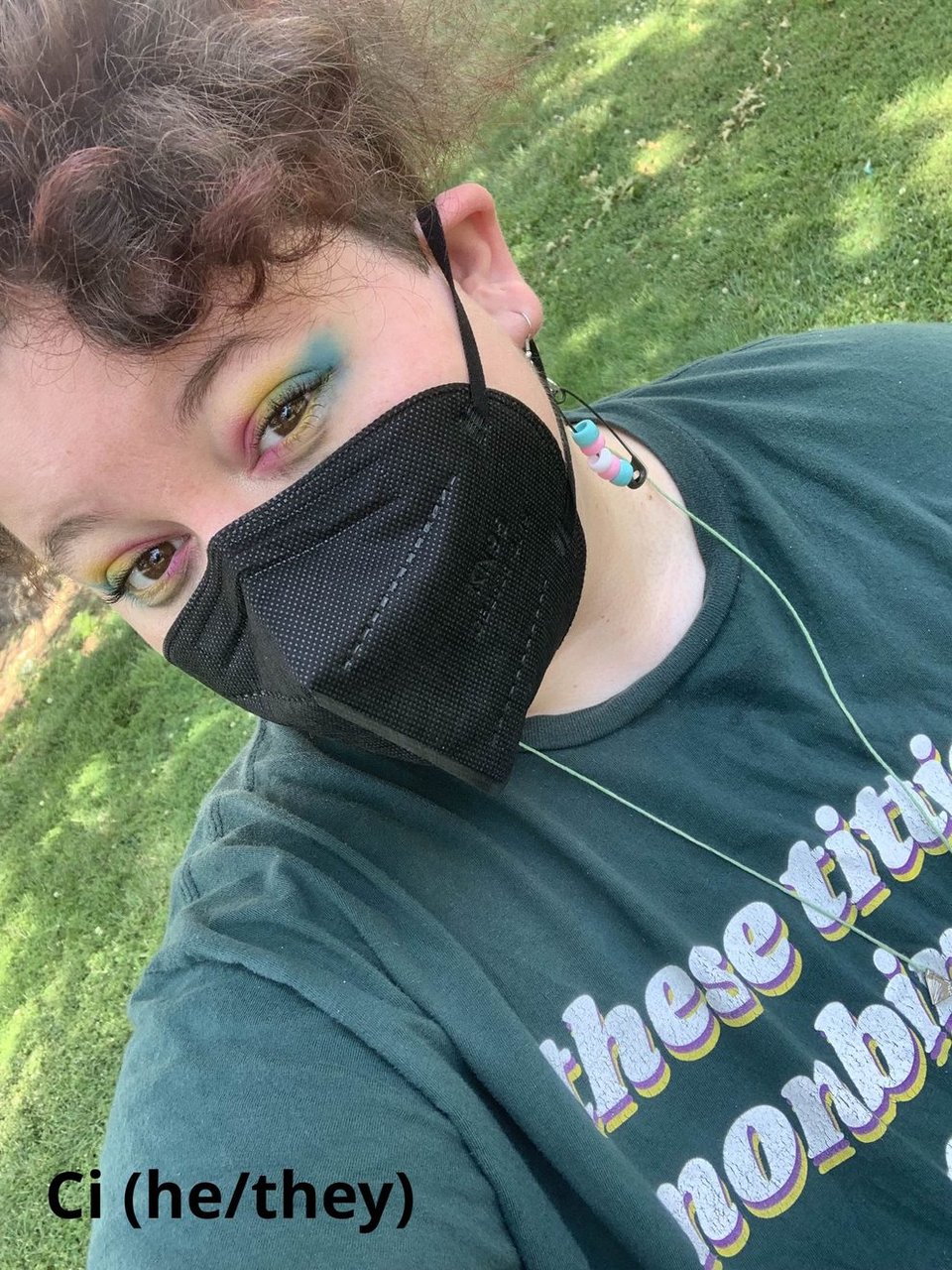 Selfie of Ci, a white fat person, is wearing a black mask with pan flag colored eyeshadow, a shirt that says "these titties nombinary too". He also has on trans safety-pin earrings.