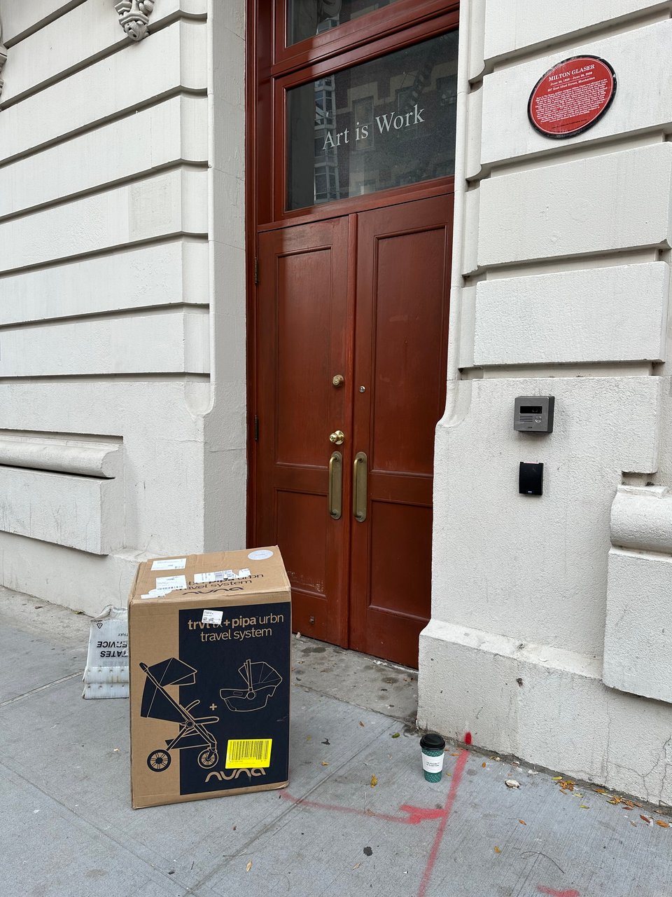 photo of exterior of Milton Glaser's studio, with a cardboard shipping box for a baby carriage in front of the door, along with a cup of coffee and some USPS bins