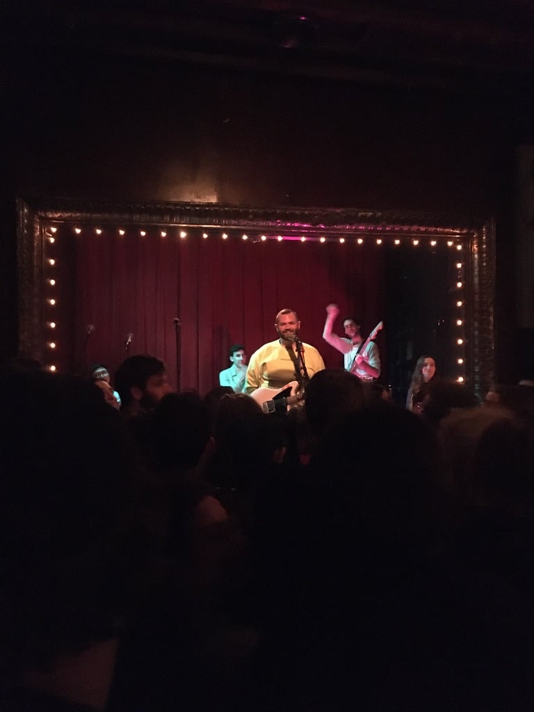 a group of people smiles and waves on a jewelbox stage above a big crowd gathered in the dark