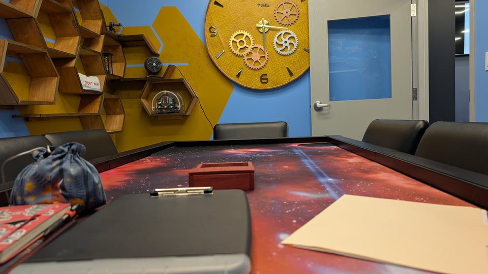 A shot of a fancy table with a topper with a cosmos print. A clipboard, some character sheets, a notebook, a bag of dice, and a wooden dice tray sit on the table, waiting for players. The walls are painted blue and contain hex-shaped, honey-comb-like shelves, and a large clock with golden, silver, and bronze gears. The door, which has a window in it, stands open.