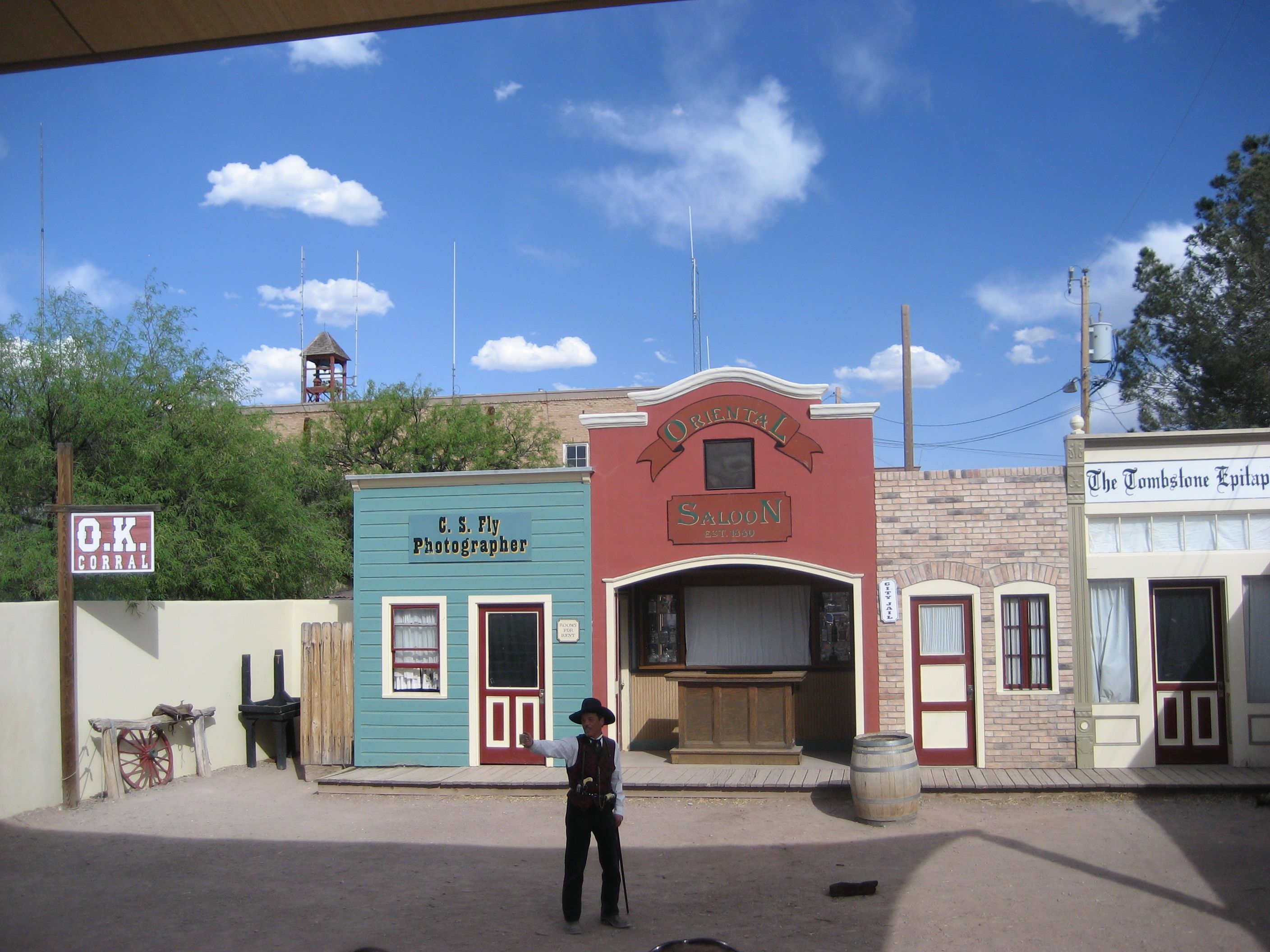 A man dressed as Doc Holliday stands in a mock up of the OK Corral with a newspaper, photographers office, and a barrel.