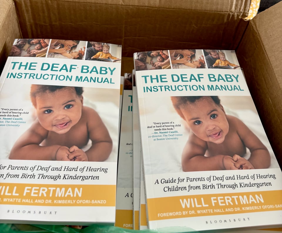 Photo of a cardboard box filled with identical books, each with a happy Black baby smiling from the cover. Title: The Deaf Baby Instruction Manual, a guide for parents of deaf and hard of hearing children from birth through kindergarten. By Will Fertman, foreward by Dr. Wyatte Hall and Dr. Kimberly Ofori-Sanzo. Bloomsbury.