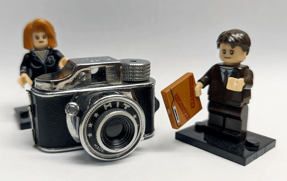 The tiny camera posed next to two Lego mini-figurines, for scale. The camera has a black body and a thick lens in chrome, and a chrome top with a viewfinder and chunky chrome film-advancer knob. The mini-figurines are male and female business people, and they are slightly taller than the camera