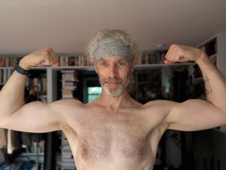 Photo of shirtless white man flexing his biceps. He’s wearing a gray headband and a black-banded Apple Watch. There are bookcases behind him.