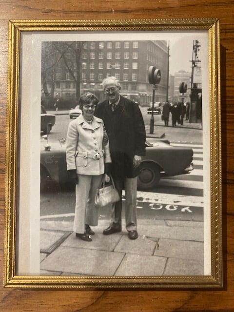 Black and white photo of a small woman in white belted jacket and matching trousers and handbag and a tall man in a short dark overcoat, standing at an intersection in London, England, in the 1970s.