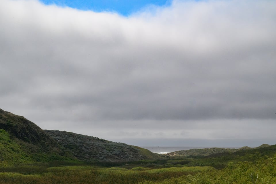 First glimpse of the ocean, between hills. The clouds are getting thicker, with only a small patch of blue visible.