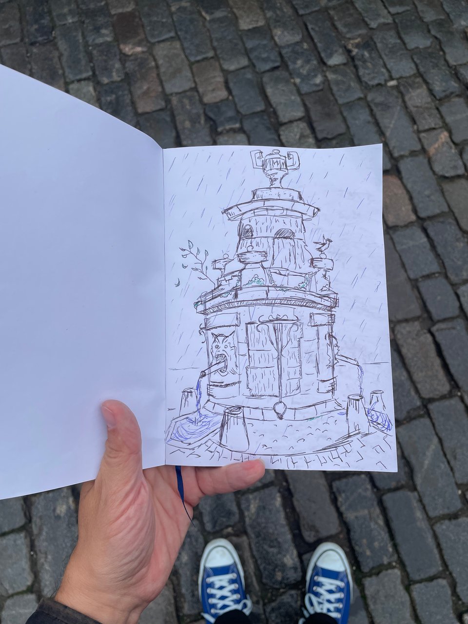 A hand holds a notebook over cobblestone street. There's a sketch of an old fountain covered in moss and demon heads.