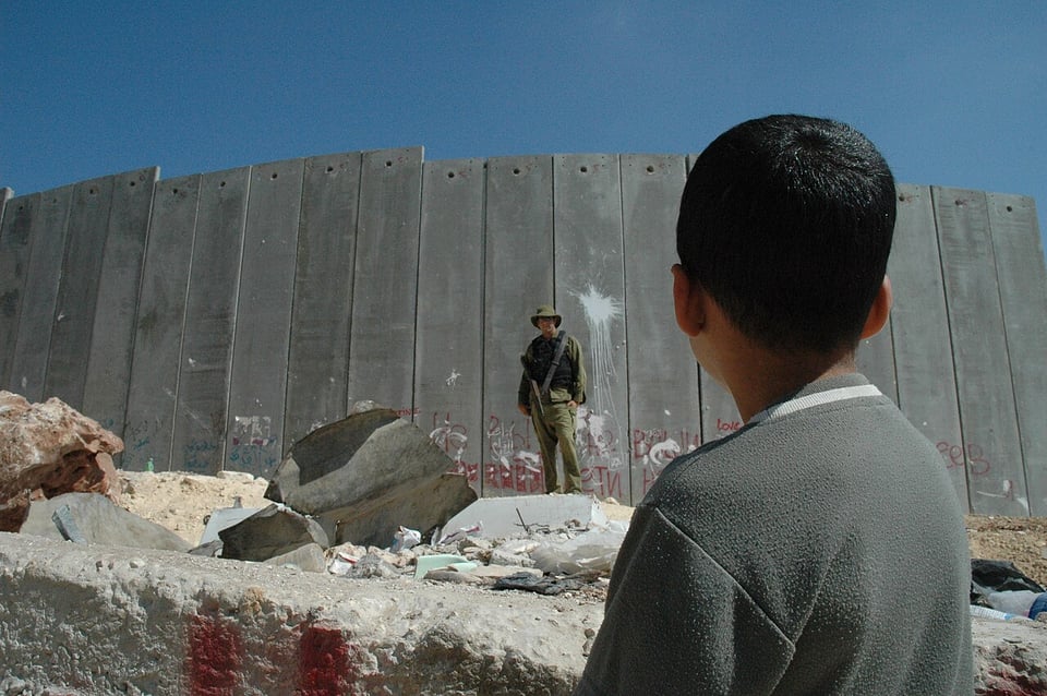 A Palestinian child looks away from the camera in the foreground, looking toward the the Israeli soldier and apartheid wall which looms over the whole photo.