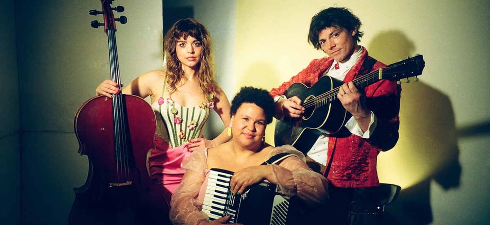 A woman with long blondish hair on the left holds a cello in her right arm. To her right, and in front of her, a woman with close-cropped dark hair sits with her arms around an accordion. To her left, a man in a red smoking jacket plays a black acoustic guitar.