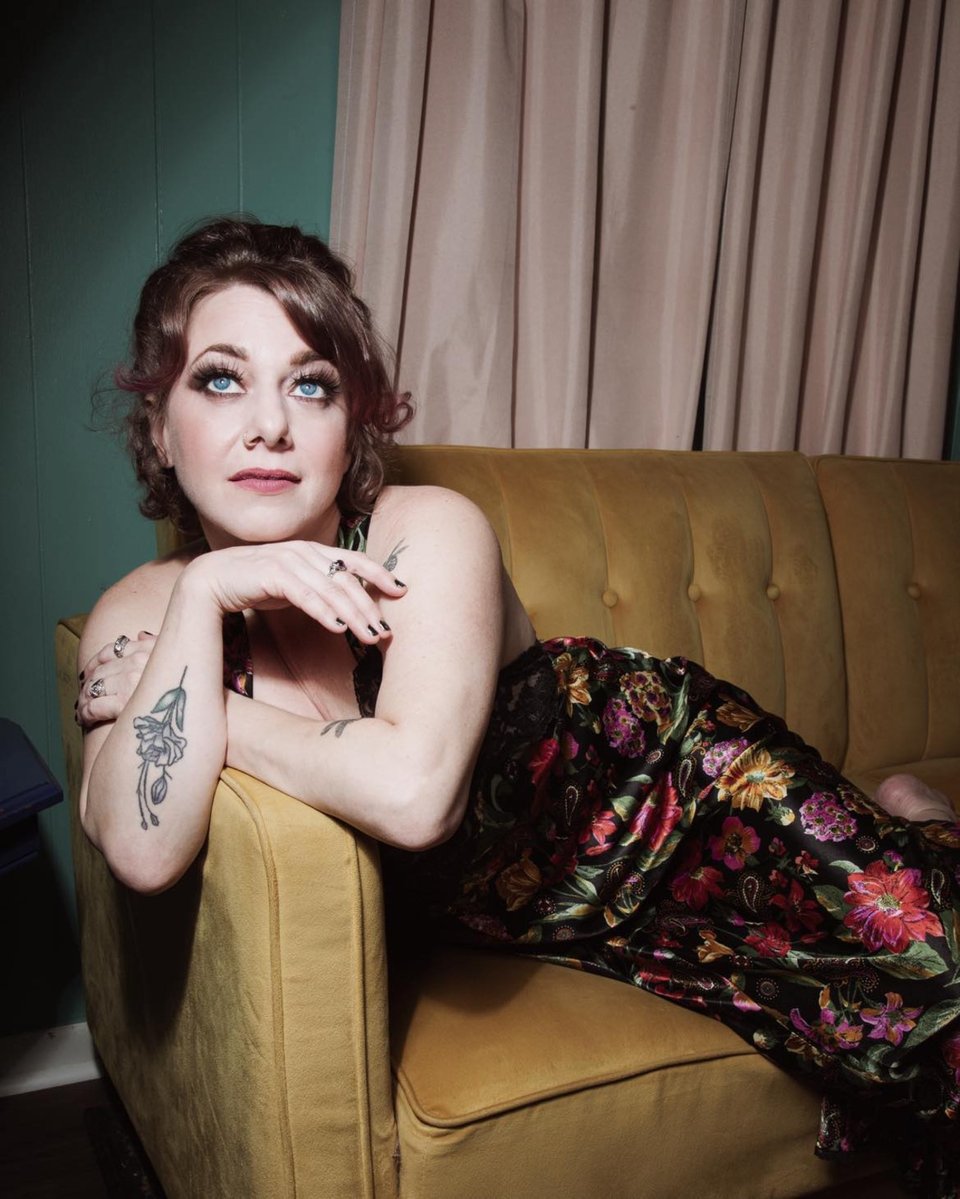 A woman in a black floral-print dress lounges on a couch with her chin resting on her right hand.