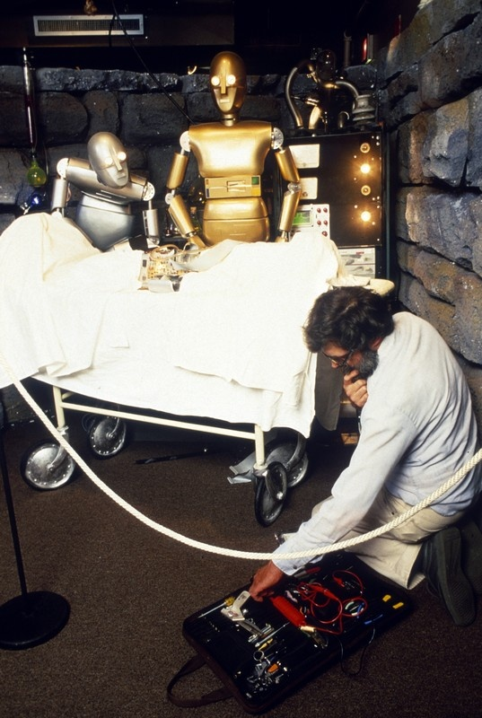 Another photograph of an employee working on setting up the robot Frankenstein scene described above. This employee is sitting on the floor with a carrying case full of electronic equipment.