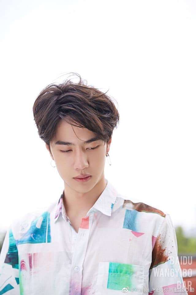 Photo of Wang Yibo with light brown, lightly tousled hair, looking down and to his left. He's wearing a white button-up with bright pastel teal-pink-green patches here and there. He's wearing two earrings; the one in his left ear isn't an earring, but a safety pin.