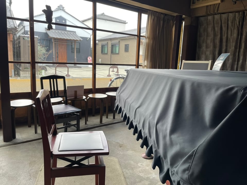 A room with a covered grand piano. Two chairs are situated near it. A large glass sliding door leading to the street is lined with chairs for spectators.