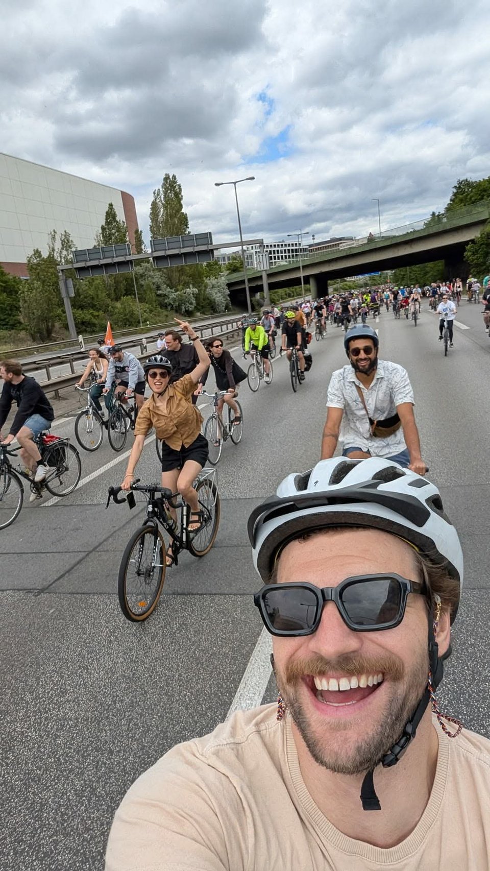 Selfie by a guy on a bike, with Jenny riding behind with a raised hand, and Dillan also, followed by a crowd of cyclists.