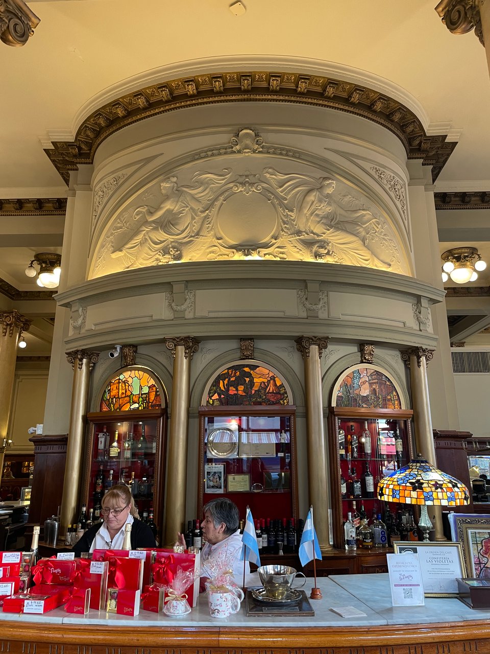 Two women work behind an elaborate cafe counter with stained glass in the displays behind them.