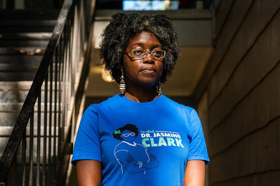 Dr. Jasmine Clark stands in a stairwell wearing glasses, patterned earrings and a bright blue campaign T-shirt that reads “re-elect Dr. Jasmine Clark.”