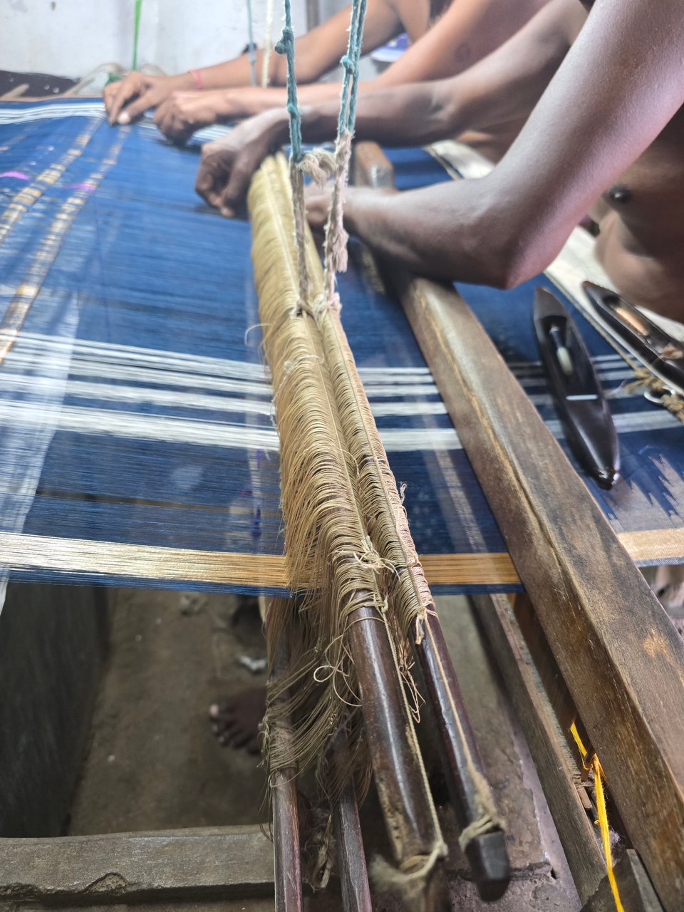 An indigo blue warp passing through two heddles and a reed. Two weavers sit side by side, tying together broken threads. Their arms can be seen next to each other as they work. In the forefront, a shuttle lies on top of the fabric, next to the gold temple and gold border.