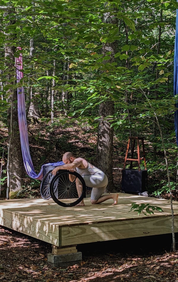 A square 10x10 foot raised platform in the woods, lit by dappled sunlight through many trees. The set objects and fabrics lean on or hang from nearby trees. I am on one knee in the center, in profile, holding a detached wheelchair wheel alongside my body, in the middle of plucking its spokes like a harp.