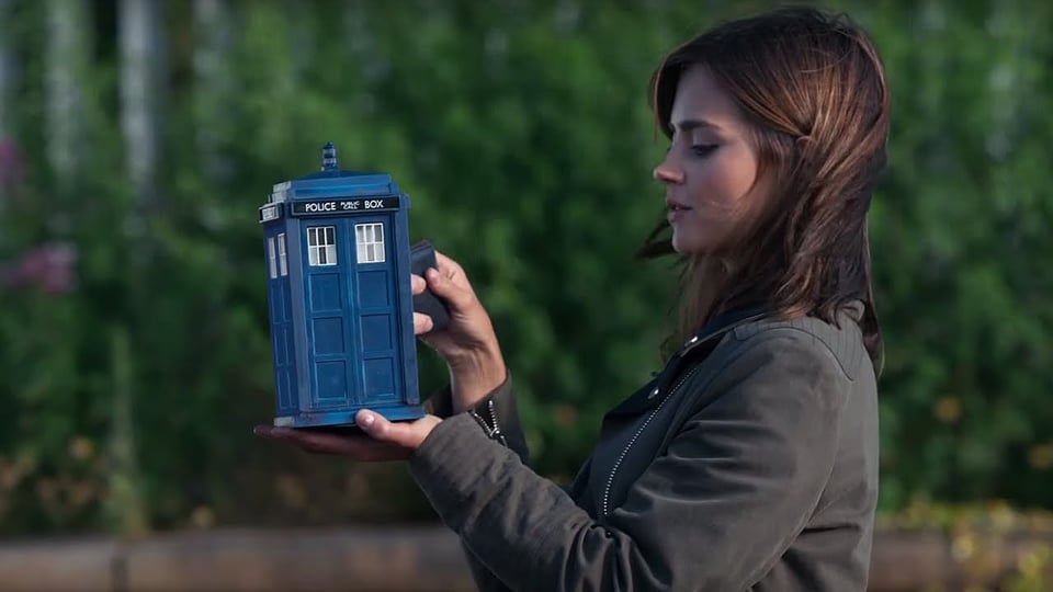 Clara, played by Jenna Coleman, holds a tiny police box in her hands. She has brown hair and is wearing a gray jacket.