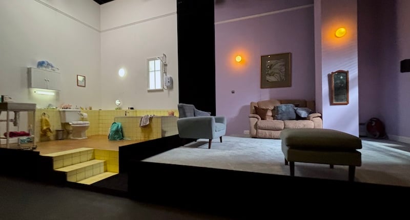 Photo of a set showing a kind-of slightly run-down but only slightly bathroom, and a lilac living room with a couple of sofas, it's very well designed and satisfying to look at
