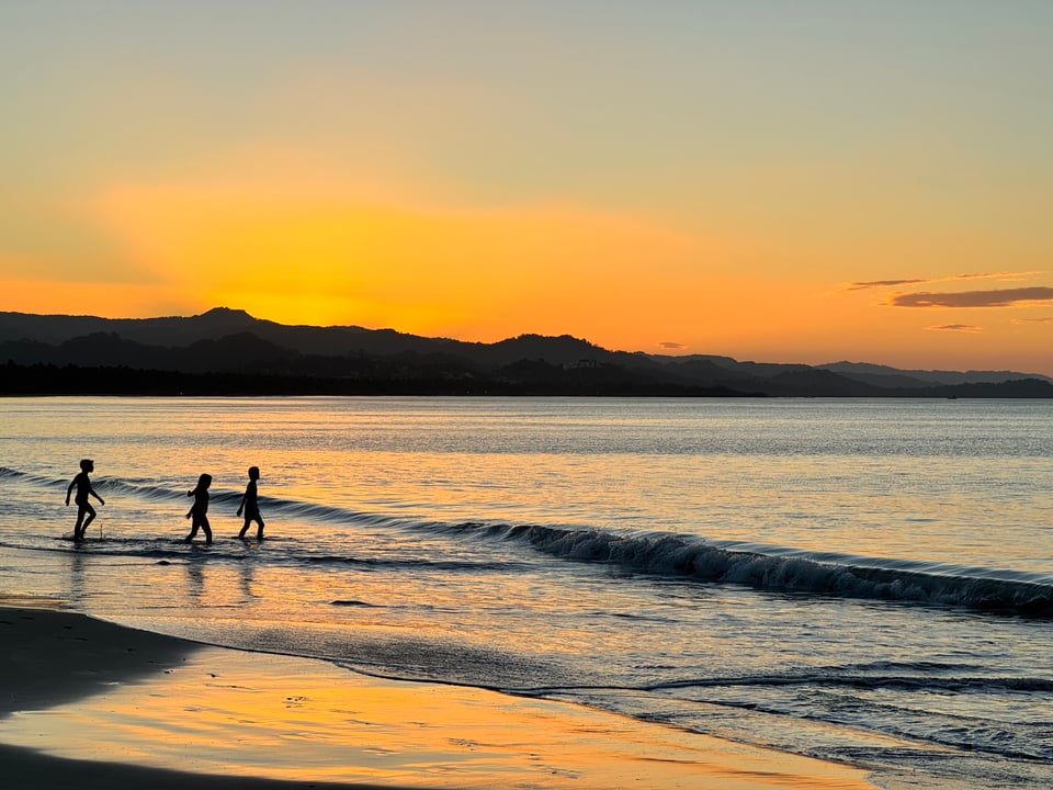 Silhouetted figures of three children walking in the shallow ocean water at sunset, with a vibrant orange and yellow sky reflecting on the wet sand and water.